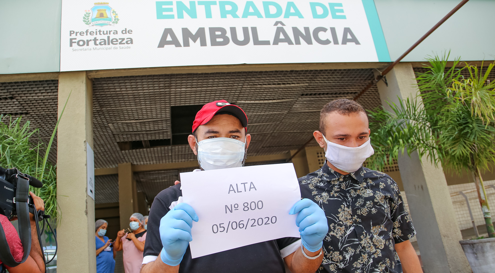 homem de máscara cirúrgica e luvas saindo de hospital do PV segurando papel que diz 'alta numero 800' e seu filho ao lado também usando máscara